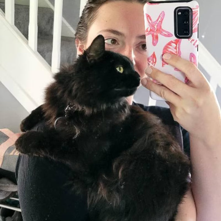 Person holding a black cat and taking a selfie with a phone case featuring starfish designs.